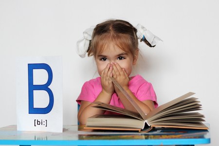 Cute little girl near B letter closing her mouth with hands in front of the open book, indoor isolated portrait over the white backgroundの写真素材