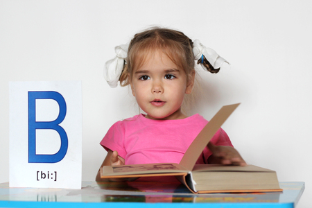 Cute little girl near B letter thinking in front of the open book, indoor isolated portrait over the white backgroundの写真素材