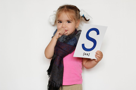 Pretty little girl in the scarf with S letter in one hand giving a cough in the fist because of cold, indoor isolated portrait over the white backgroundの写真素材