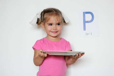 Pretty little girl with P letter smiling and holding a white plate, indoor isolated portrait over the white backgroundの写真素材