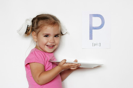 Pretty little girl with P letter smiling and holding a white plate, indoor isolated portrait over the white backgroundの写真素材