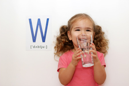 Pretty small girl with W letter on white background drinking a glass of water in her hands, indoor portraitの写真素材