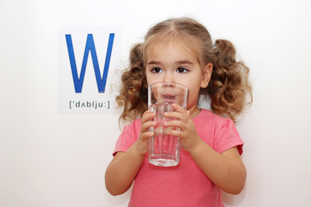 Pretty small girl with W letter on white background drinking a glass of water in her hands, indoor portraitの写真素材