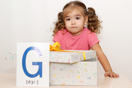 Pretty small girl with fun face shrugging her shoulders near the gift box with G letter on it, indoor portraitの写真素材