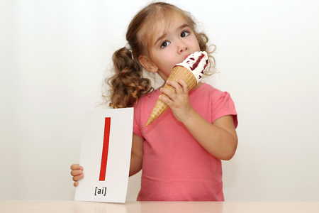 Pretty small girl eating an ice-cream on the white background with I letter on it, indoor portraitの写真素材