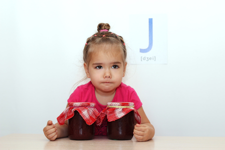 Pretty small girl hugging glass jars with jam on white background with J letter on it, indoor portraitの写真素材