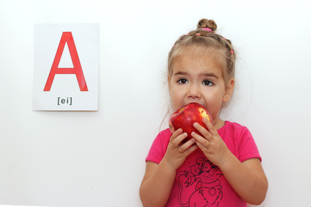 Cute little girl eating a red apple, indoor isolated portrait over the white background with A letter on itの写真素材