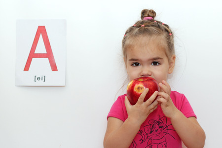 Cute little girl eating a red apple, indoor isolated portrait over the white background with A letter on itの写真素材