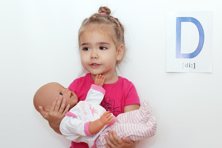 Pretty small girl hugging a doll and singing lullaby to it over white background with D letter on it, indoor portraitの写真素材