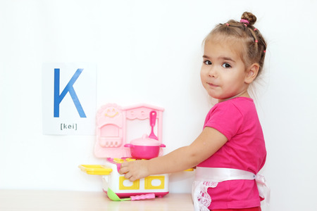 Pretty small girl playing with a toy kitchen over white background with D letter on it, indoor portraitの写真素材