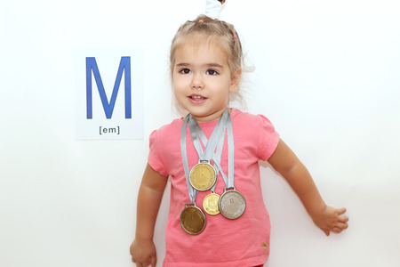 Pretty small girl with several medals on her neck over white background with M letter on it, indoor portraitの写真素材