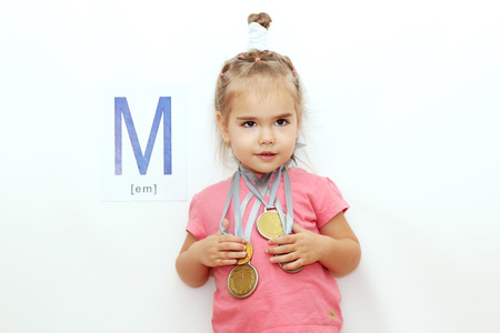 Pretty small girl with several medals on her neck over white background with M letter on it, indoor portraitの写真素材