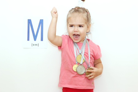 Pretty small girl with several medals on her neck over white background with M letter on it, indoor portraitの写真素材
