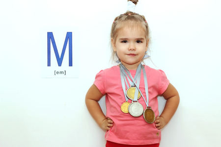 Pretty small girl with several medals on her neck over white background with M letter on it, indoor portraitの写真素材