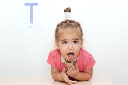 Pretty small girl wearied a tie sticking her tongue out over white background with T letter on it, indoor portraitの写真素材
