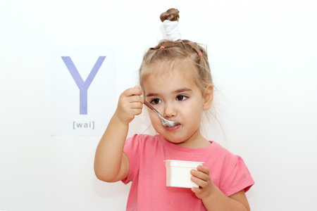 Pretty small girl eating a yogurt over white background with Y letter on it, indoor portraitの写真素材