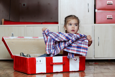 Cute happy little girl sitting in the red and white fascinated vintage suitcase and preparing to travel, indoor portraitの写真素材