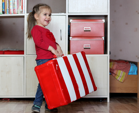 Cute happy little girl lifting red and white fascinated vintage suitcase and carry it, indoor portraitの写真素材