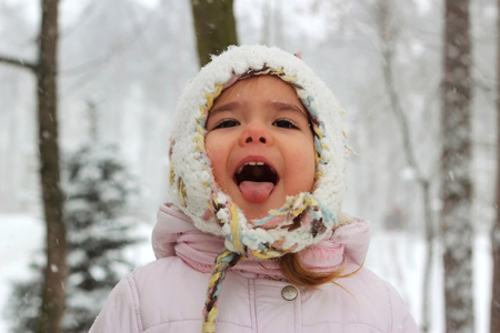 Little toddler girl in warm coat and knitted hat having fun in the winter forest and trying to catch snowflakes with her mouth, outdoor portraitの写真素材