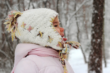 Little toddler girl in warm coat and knitted hat having fun in the winter forest and trying to catch snowflakes with her mouth, outdoor portraitの写真素材