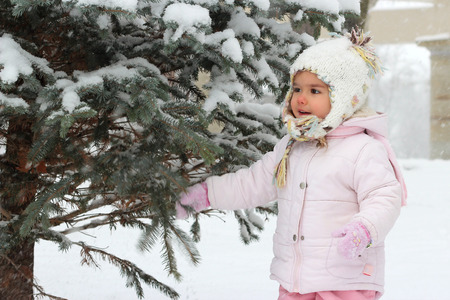 Happy toddler girl in warm coat and knitted hat shaking snow off the tree and having a fun in the winter forest, outdoor portraitの写真素材