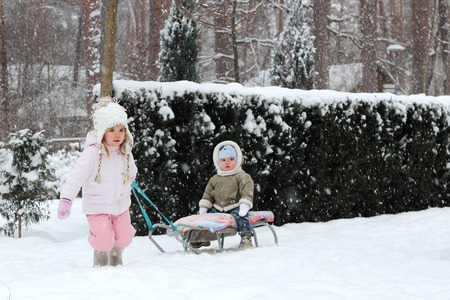 Little toddler girl in warm coat and knitted hat pulls her happy toddler brother sitting on the sledge in the winter forest, outdoor portraitの写真素材