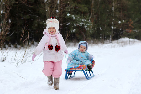 Little toddler girl in warm coat and knitted hat pulls her happy toddler brother sitting on the sledge in the winter forest, outdoor portraitの写真素材
