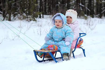 Little toddler girl in warm coat and knitted hat and happy toddler boy sitting on the sledge in the winter forest, outdoor portraitの写真素材