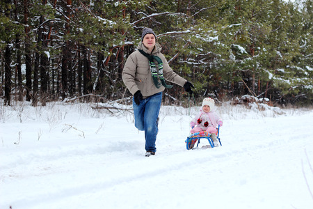 Happy father running with sledges on a winter day, little toddler girl in warm coat and knitted hat sitting on the sledge, family weekend outdoorの写真素材