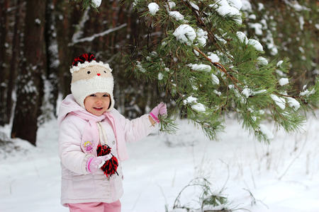 Happy toddler girl in warm coat and knitted hat shaking snow off the tree and having a fun in the winter forest on vacation, spending time outdoorsの写真素材