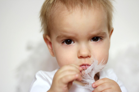 Portrait of tender blond toddler in a white t-shirt touching himself with white feather over white background, romantic and love conceptの写真素材