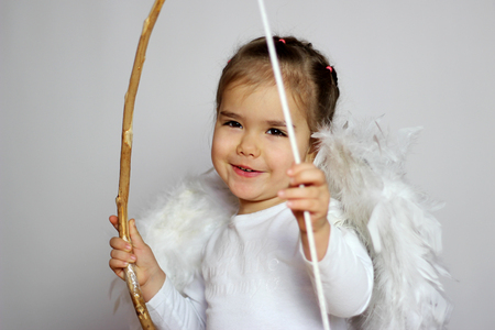 Closeup portrait of cute toddler girl in a white t-shirt with white wings as a Cupid looking infused and inspired with bow in her hand over the white background, Saint Valentines Day card, romantic and love conceptの写真素材