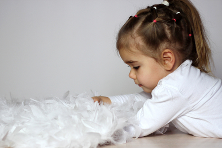 Portrait of cute toddler girl in a white t-shirt with white wings as a Cupid lies putting her arms under her wings over white background, Saint Valentines Day card, romantic and love conceptの写真素材
