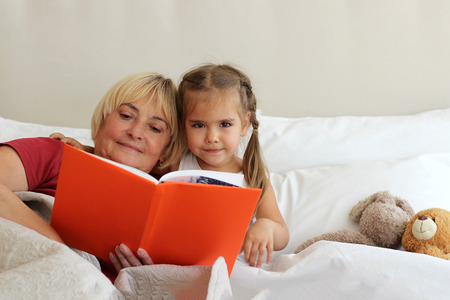 Happy small girl lying in bed with her grandmother who is reading a book with fairytales at home, bedtime, childhood and family concept, close-up indoor portraitの写真素材