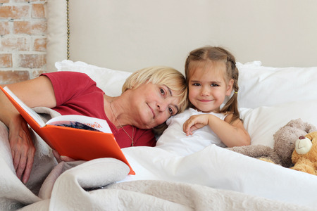 Happy small girl lying in bed with her grandmother who is reading a book with fairytale at home, bedtime, childhood and family concept, close-up indoor portraitの写真素材