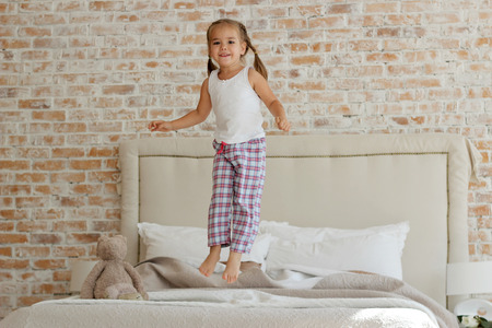 Cute happy little girl in pajama jumping on her parents white bed in the morning at home, childhood and family concept, horizontal indoor portraitの写真素材