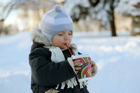 Portrait of handsome toddler boy in winter coat, knitted hats, and scarf holding paper mug and drinking cocoa in the snow-covered park in winter day, happy family concept, child playing outdoorsの写真素材
