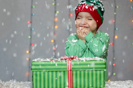 Portrait of happy toddler boy in a green hat like a little elf with gift box on the snow over white Christmas background, winter holiday concept, happy family and childhoodの写真素材