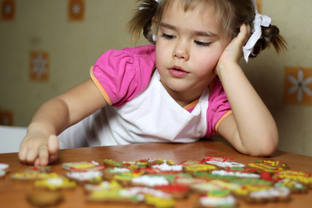 Portrait of pretty little girl in white apron dreaming about a gift from Santa Claus near colorful cookies she baked and drawn for him by herself, happy family concept, winter holidayの写真素材