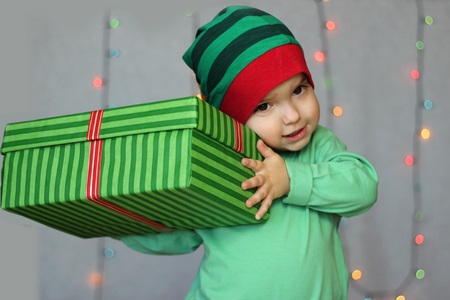 Portrait of happy little boy like elf holding a box with gift over Christmas background, winter holiday family conceptの写真素材