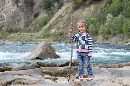 Pretty small girl with two braids and wood stick standing on the bank of the mountain river in the Carpathians on a warm day, travel and active lifestyle, outdoorの写真素材
