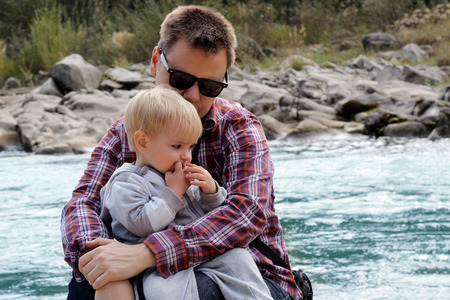 Serious father in black sun eyeglasses and checkered shirt hugs his handsome toddler boy looking on the same direction to see beautiful nature, outdoorの写真素材