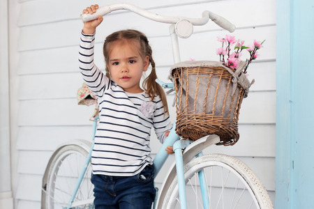 Pretty small girl in jeans and striped t-shirt stay near the vintage white bicycle with basket with flowers over white wooden wall and dream, beauty and fashion concept, indoor portraitの写真素材