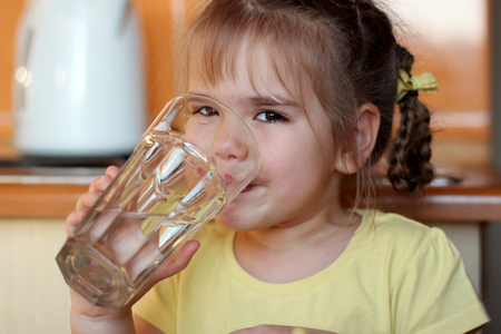 Pretty little girl drinking water from a glass in her hands in the kitchen at home, food and drink concept, indoor close-up portraitの写真素材