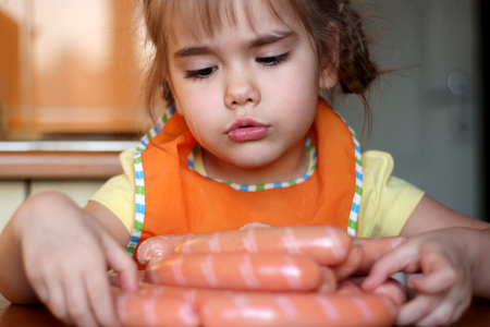 Cute preschooler girl looking at a heap of sausages with regret in the kitchen at home, indoor portrait, food and drink concept の写真素材