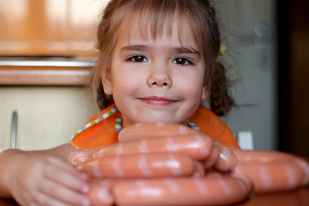 Cute preschooler girl hugging a heap of sausages like beads in the kitchen at home, indoor portrait, food and drink concept の写真素材