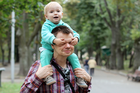 Smiling little boy closes his fatherâs eyes while he carrying his son on the shoulders, weekend time together, happy family concept, outdoor portraitの写真素材
