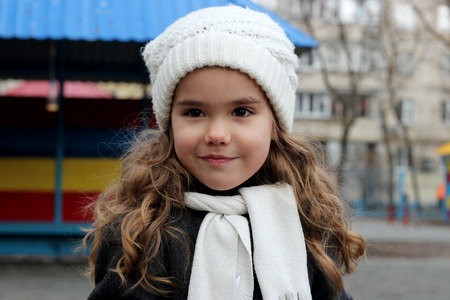 Cute little girl with curly hairs in hat staying at playground in the kindergarten at first sunny spring day and looking cheerfully, happy family weekend, outdoor closeup portraitの写真素材