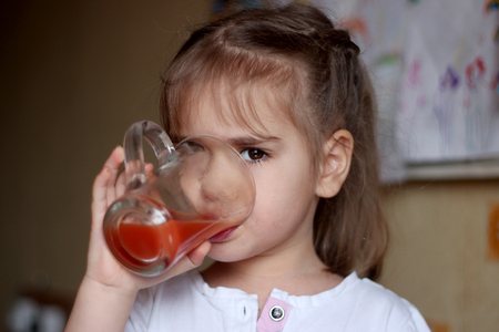 Portrait of gorgeous little girl drinking a glass of tomato juice at home, food and drink concept, healthy food, indoorの写真素材