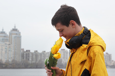 Young handsome teenage boy in yellow coat with ear-phones smelling a bunch of yellow tulips and waiting for a date on the bank of the river in rainy day in a big city, outdoor portraitの写真素材